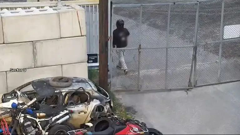 En person med hjelm og mørke klær går gjennom en åpen metallport, med en havarert bil og annet rusk synlig til venstre for porten, likt åstedet hvor en rask reaksjon hindrer tyveri - bevæpnet mann pågrepet hos bilopphugger i Arendal.